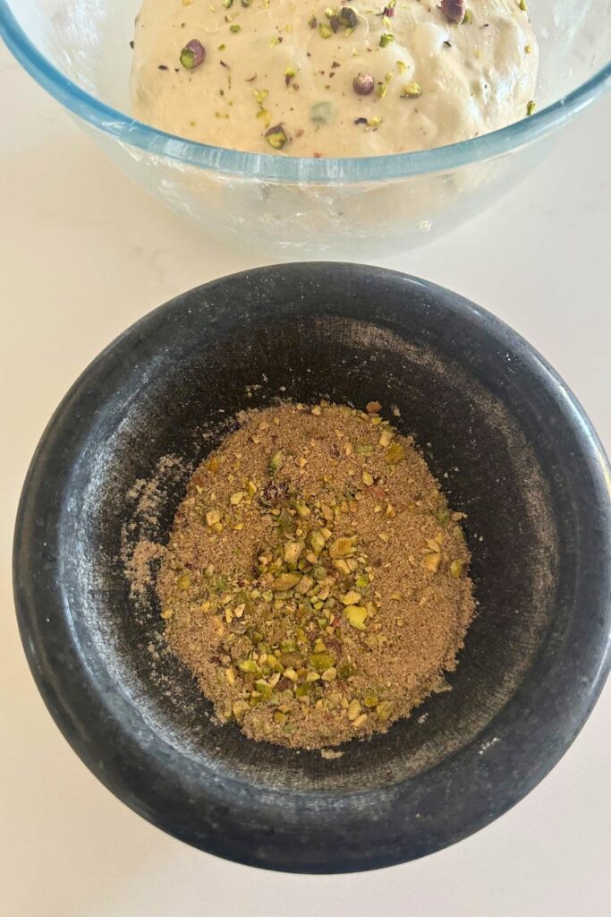 A black granite dish containing the pistachio and spice mixture to add to the middle of the sourdough during the shaping process. You can  see the dough bulk fermenting in the background of the bowl.