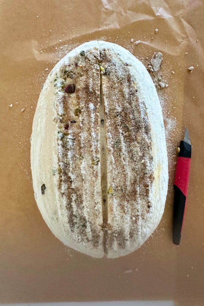 A loaf of sourdough pistachio bread that has been scored and is ready to bake. There is a small red handled bread lame in the photo too.