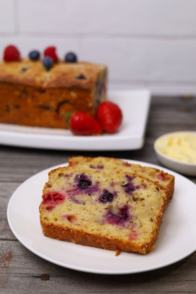 A feature image of sourdough berry banana bread. There are two slices of sourdough berry banana bread displayed on a white plate. You can see the rest of the loaf in the background of the photo garnished with fresh berries.