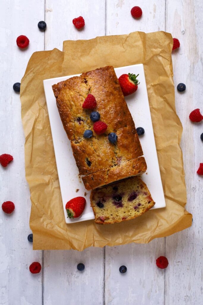Photo of sourdough berry banana bread loaf taken from above. There are a few slices cut from the loaf and laid over so you can see the bright berries inside. There are also fresh berries garnishing the top of the quick bread.