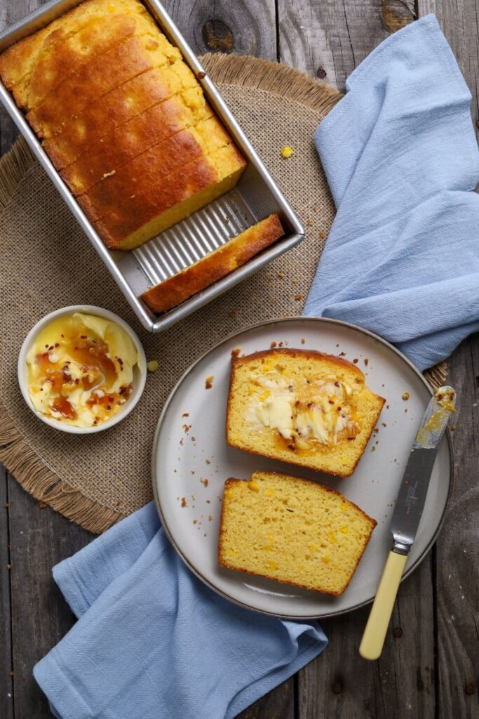 A loaf of sourdough cornbread sitting on a woven placemat. Some of the sourdough cornbread has been sliced and is on a white plate, drizzled with some butter and hot honey. There is a pale blue linen dish towel decorating the scene.