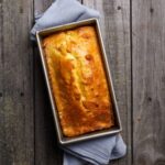 A sourdough cornbread loaf baked in a silver loaf pan. There is a pair of pale blue napkins decorating the loaf pan.