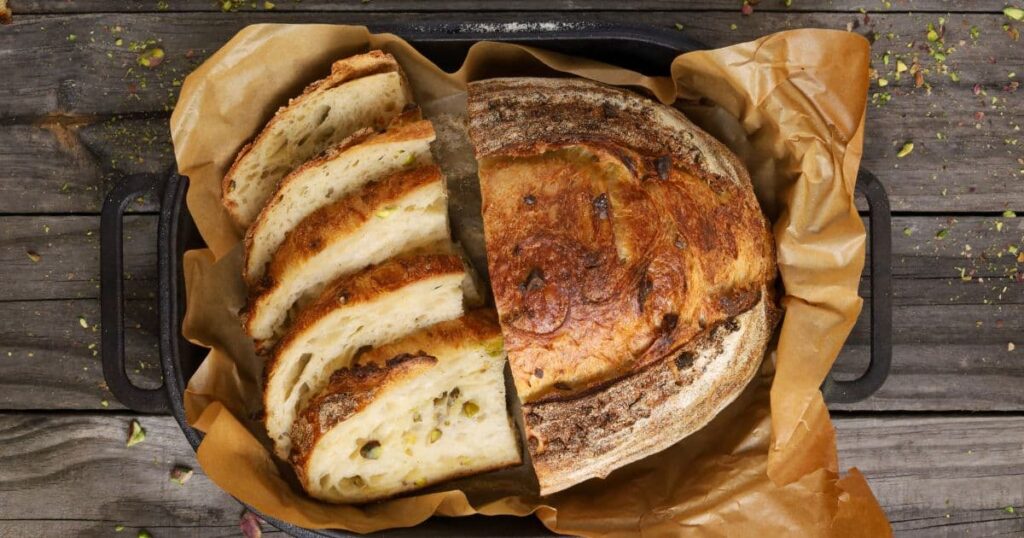 A sourdough croissant loaf crossed with a sourdough pistachio loaf. Half of the loaf is sliced so you can see the crumb. The whole loaf is displayed in a black cast iron bread pan.