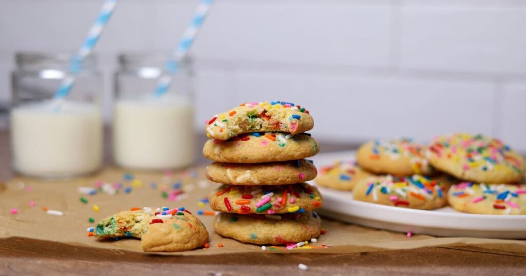 A stack of 5 sourdough funfetti cookies. The top cookie has a bite taken out of it, as does the cookie sitting next to the stack. There is a glass of milk with a blue straw in the background of the photo.