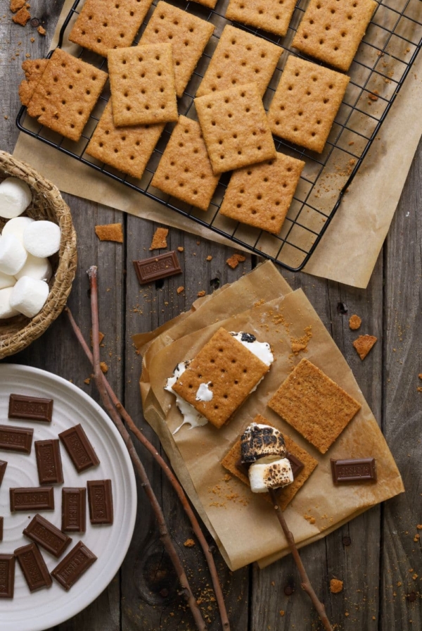 A photo taken from above showing a black wire rack of sourdough graham crackers at the top of the photo. There is also a small basket of white marshmallows and a white plate of Hershey's pieces. To the right of the photo there is a piece of parchment paper where some s'mores have been made with toasted marshmallows and sticks.