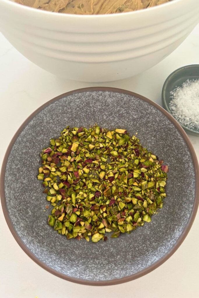 A grey stoneware bowl of roughly chopped purple and green pistachios. There is a also a small bowl of flaky sea salt.