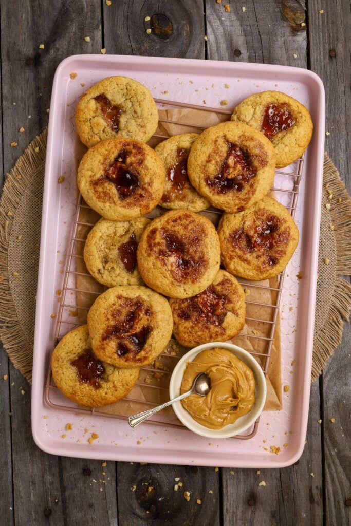Flat lay of a bunch of sourdough peanut butter and jelly cookies layered on a pink baking tray. There is a bowl of peanut butter on the tray too.
