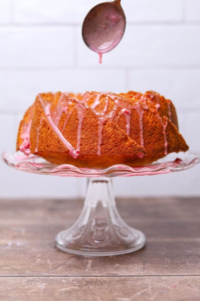 A sourdough pound cake that has been baked in a bundt pan and then displayed on a glass cake stand. The cake is drizzled in a raspberry glaze and you can see the spoon drizzling the glaze in the top of the photo.