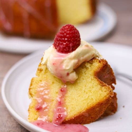 A slice of sourdough pound cake on a white plate. The yellow cake slice is topped with whipped cream and drizzled with pink raspberry glaze. A fresh raspberry sits atop the whipped cream. You can see the bottom of the rest of the sourdough pound bundt cake in the background of the photo.
