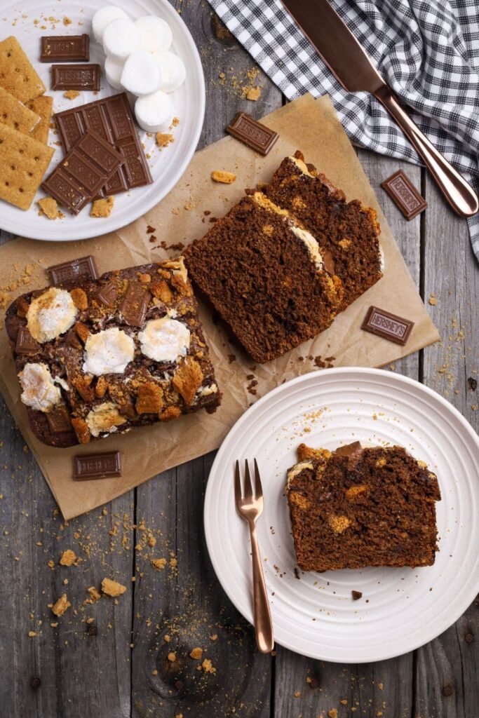 A flat lay photo of sourdough s'mores banana bread taken from above. There is a loaf of sourdough s'mores banana bread that has been sliced up so you can see the crumb inside. A piece of the loaf is sitting on a white plate.