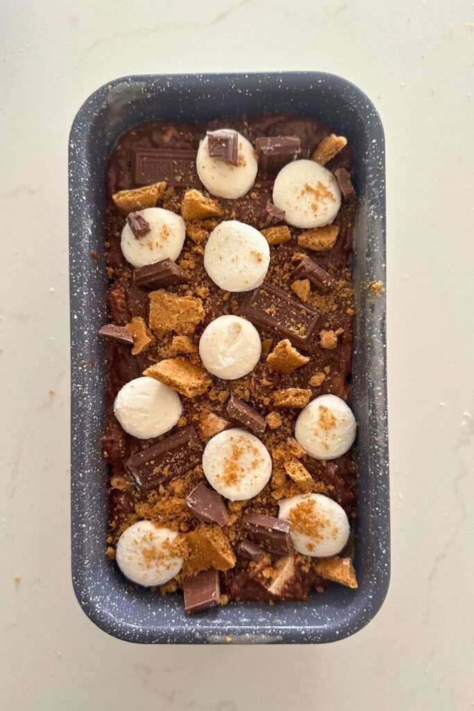 A grey colored loaf pan filled with sourdough s'mores banana bread topped with marshmallows, crushed graham crackers and Hershey's pieces.