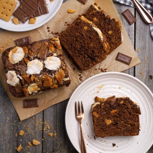 A flat lay photo of sourdough s'mores banana bread taken from above. There is a loaf of sourdough s'mores banana bread that has been sliced up so you can see the crumb inside. A piece of the loaf is sitting on a white plate.