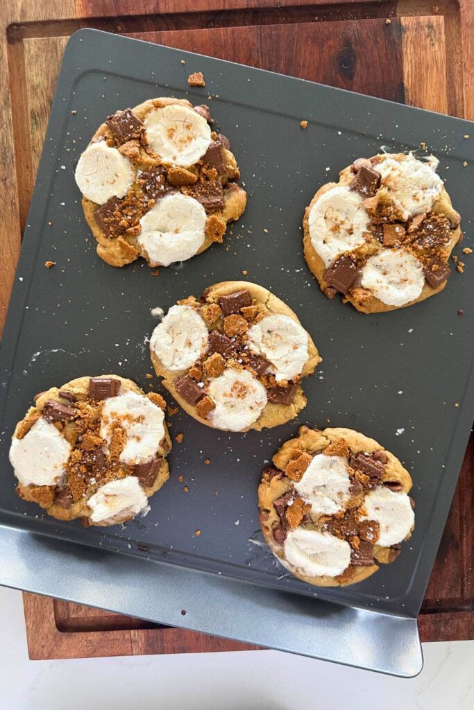5 sourdough s'mores cookies that have just been pulled out of the oven. The cookies are sitting on a baking sheet that is a top a large wooden board.