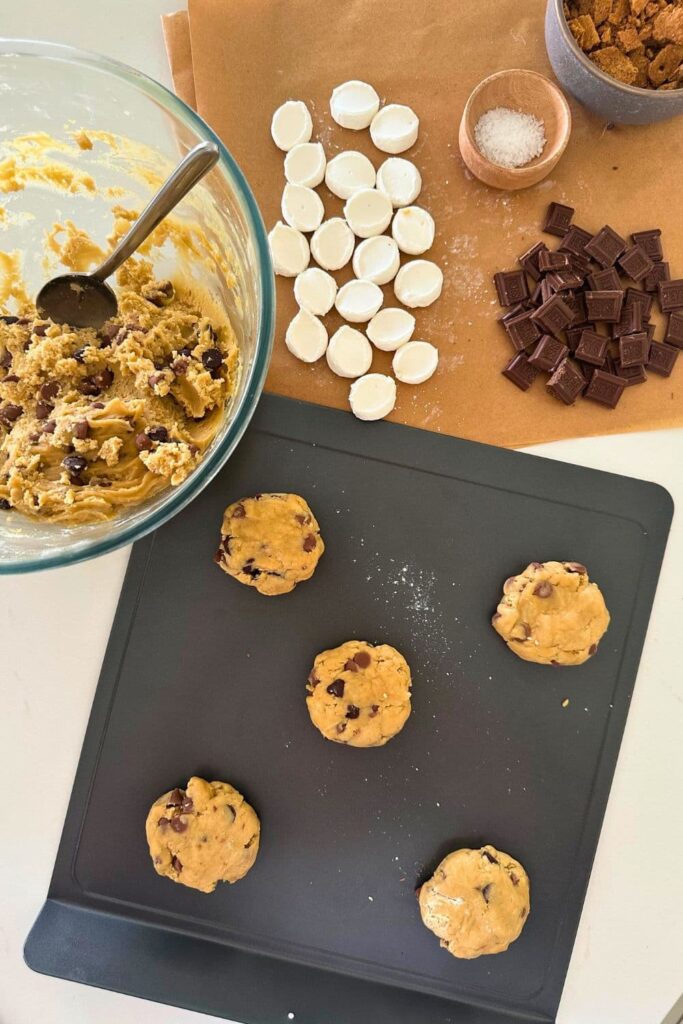 A photo taken overhead showing the process of making the cookie dough balls. You can see 5 cookie dough balls sitting on a cookie tray. Around the tray is the bowl of sourdough chocolate chip cookie dough, as well as the s'mores toppings including marshmallows, Hershey's pieces, graham cracker crumbs and a small bowl of flaky sea salt.