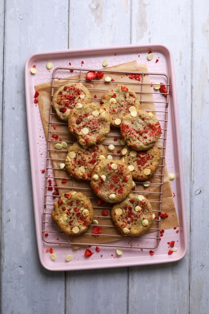 A flat lay image taken from above. There is a pink tray with a pink cooling rack sitting on top. There is a pile of sourdough strawberries and cream cookies sitting inside the tray.
