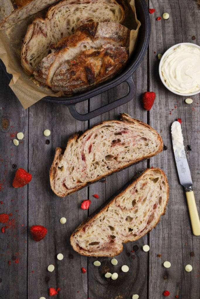2 slices of strawberries and cream sourdough bread laid on a wooden board. There are freeze dried strawberries and white chocolate chips surrounding the loaf, as well as a dish of marscapone cheese and a small knife.