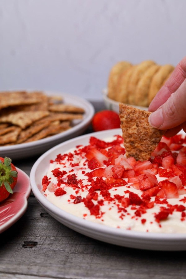 Strawberry cheesecake dip garnished with fresh and freeze dried red strawberries served on a white plate. There is a hand dipping a sourdough cracker into the dip.