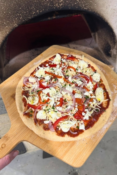 A whole wheat sourdough pizza base topped with cheese and vegetables being put into a wood fired pizza oven using a wooden pizza peel.