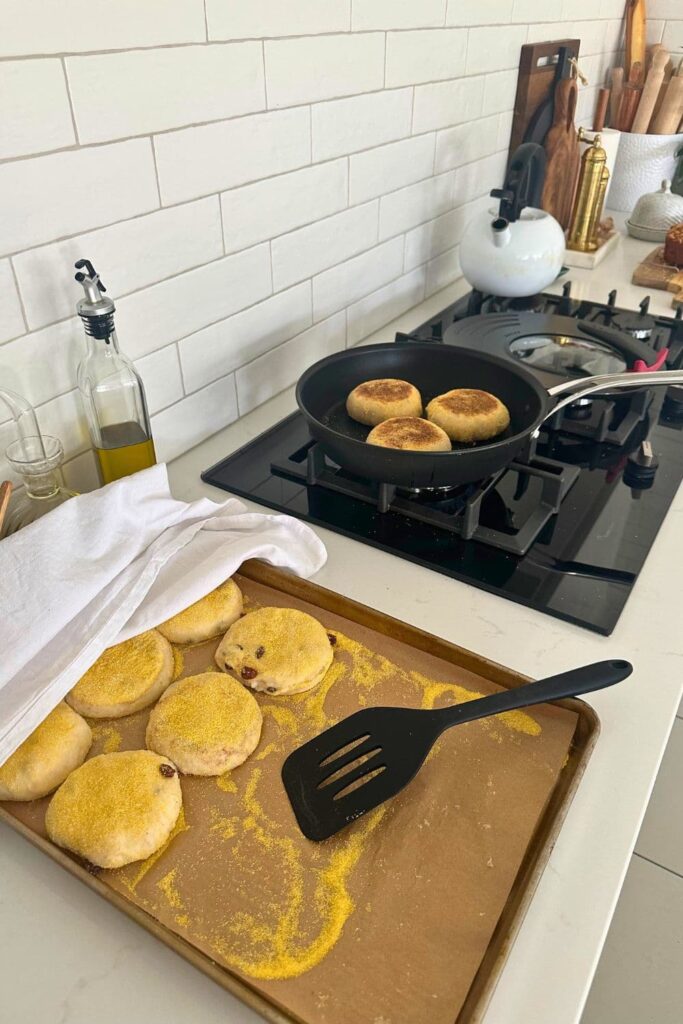 An image of a country kitchen with a hot skillet on the range. Inside the skillet are 3 sourdough cinnamon raisin English muffins baking. Next to the range is a large tray of sourdough cinnamon raisin English muffins that still need to be baked.
