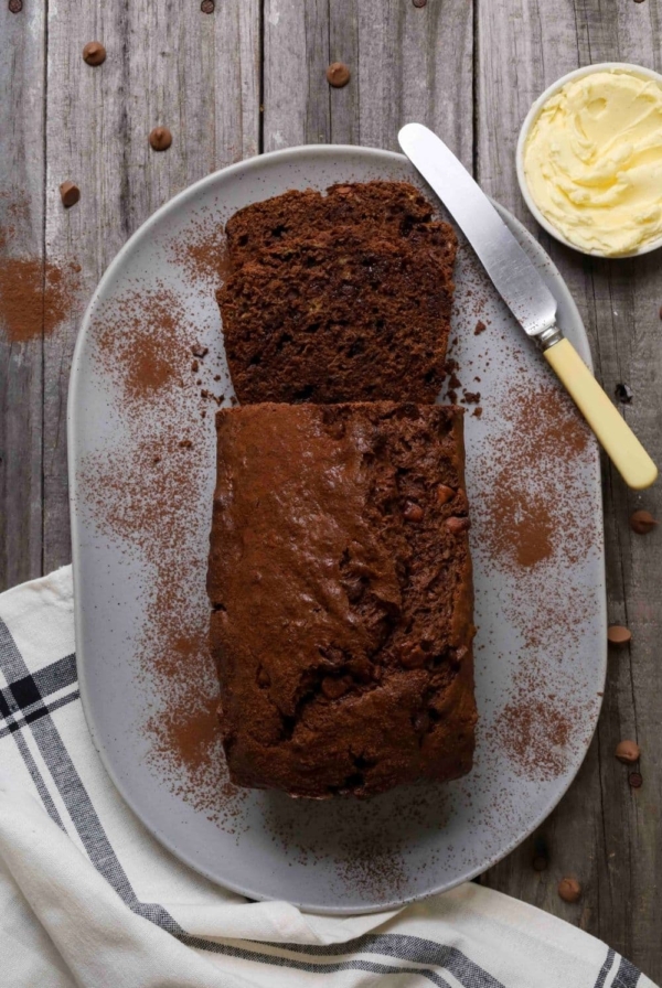A loaf of double chocolate sourdough banana bread on a light gray plate, lightly dusted with cocoa powder, with one slice cut to show the. A butter knife with a yellow handle and a small bowl of softened butter sit beside the loaf on a rustic wooden surface, with a black-and-white striped cloth partially visible underneath.