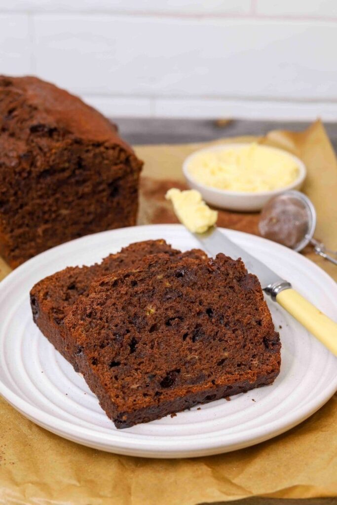 Two slices of double chocolate sourdough banana bread on a white plate, with a butter knife and bowl of soft butter in the background.