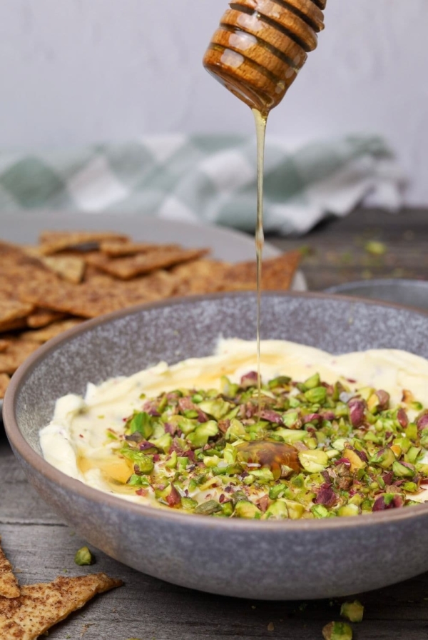 Honey pistachio cream cheese in serving bowl with honey being poured on top and a serving tray with cinnamon sugar crackers in background.