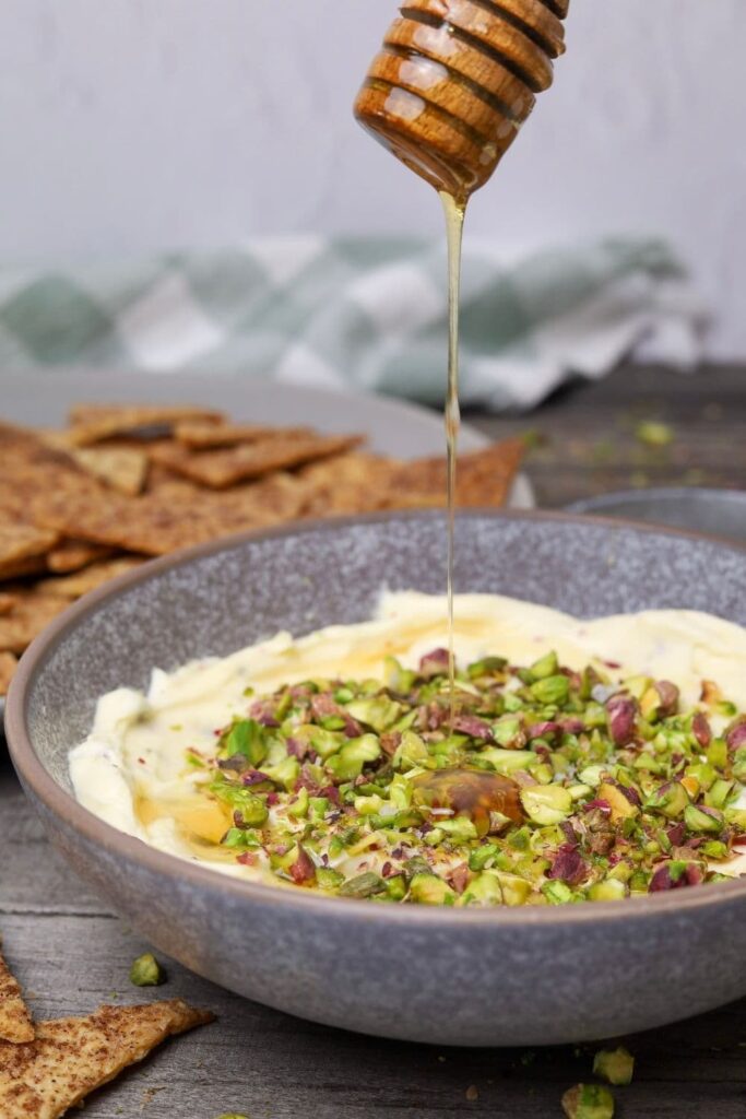Honey pistachio cream cheese in serving bowl with honey being poured on top and a serving tray with cinnamon sugar crackers in background.