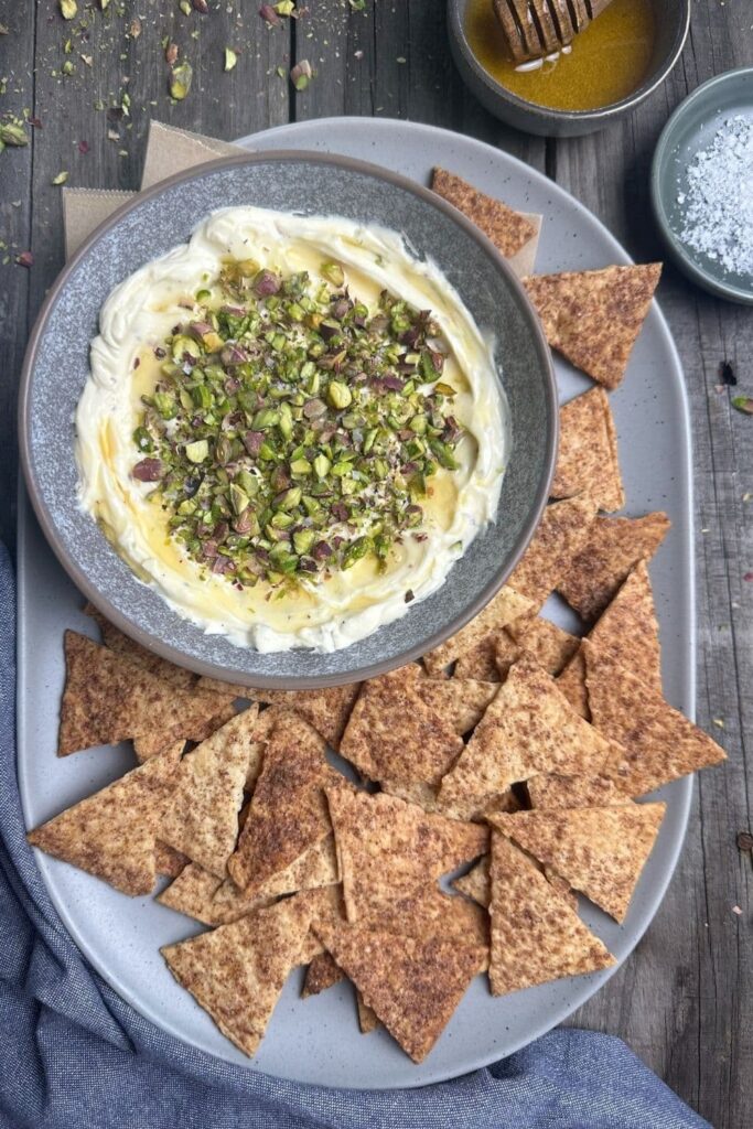 Honey pistachio cream cheese in serving bowl on a serving tray next to cinnamon sugar crackers with a bowl of honey and flaky salt in background.