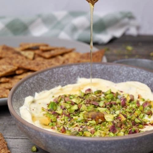 Zoom in of honey pistachio cream cheese in serving bowl with honey being poured on top and a serving tray with cinnamon sugar crackers in background.