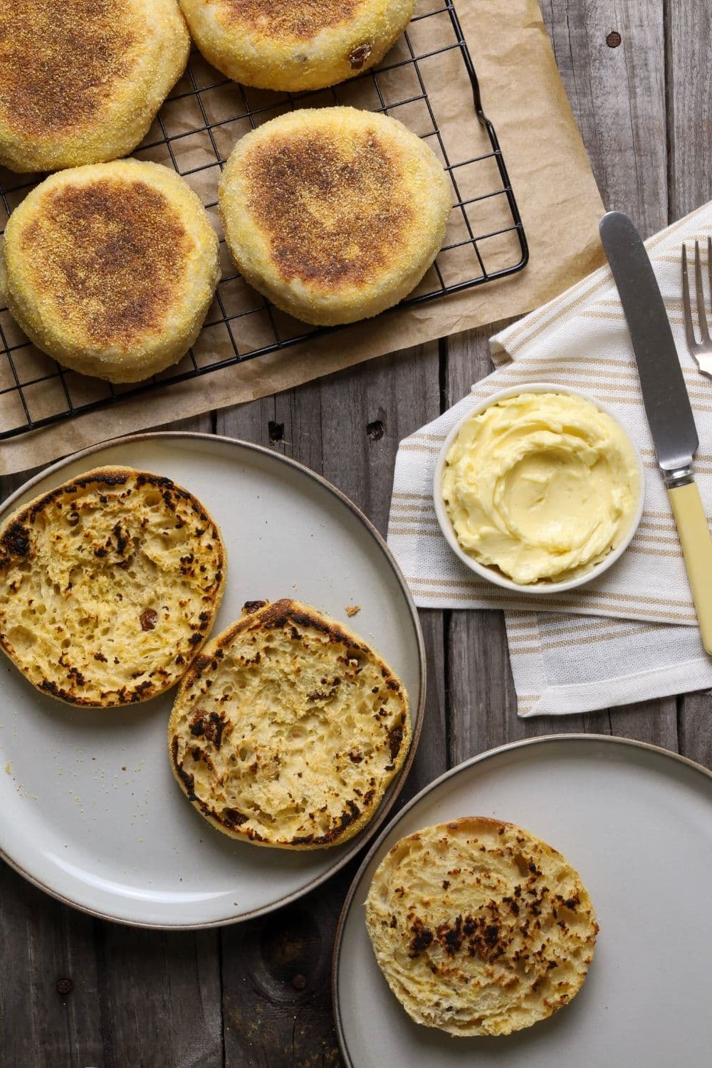 Sourdough discard cinnamon raisin English muffins on cooling rack over parchment paper next to sliced English muffins with a bowl of butter.