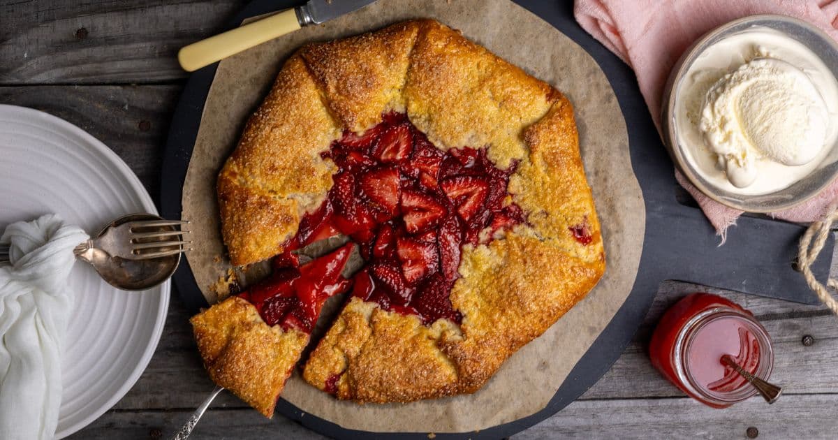 A vertical photo of a sourdough discard strawberry galette brimming with red strawberries. A slice has been taken out of the galette, but is still sitting on the plate. There is a bowl of vanilla ice cream sitting on a pink linen napkin above the galette and a small jar of strawberry sauce.