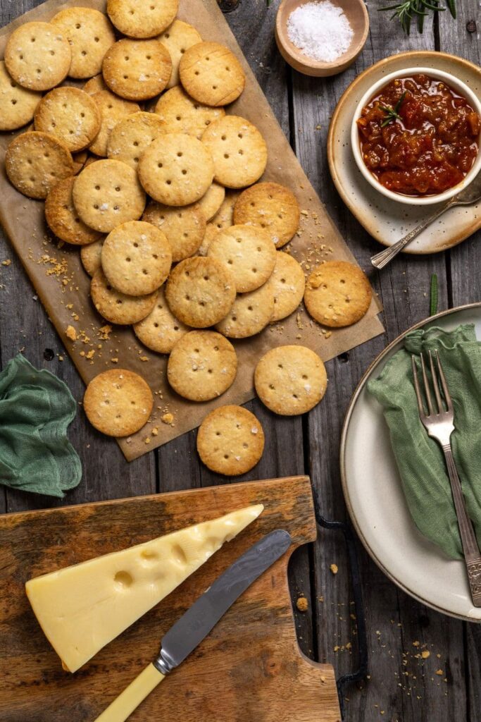 A wooden board of sourdough ritz crackers displayed with a wedge of cheese and a small dish of tomato chutney.