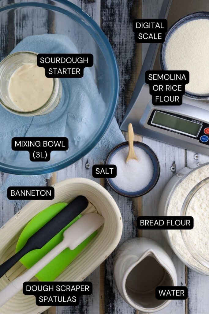 A flat lay of ingredients and equipment necessary to make sourdough bread recipe. This includes a sourdough starter, digital kitchen scale, bread flour, water, salt, banneton and spatulas.