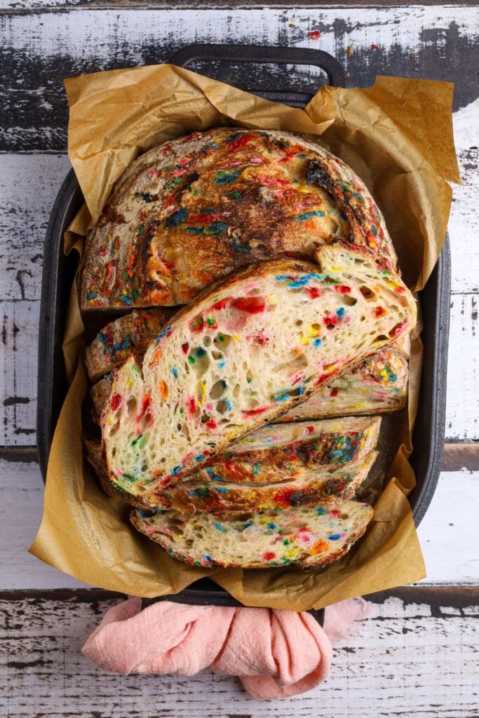 A loaf of sourdough funfetti bread sitting inside a cast iron bread pan. There is a slice of the loaf sitting on top so you can see the brightly colored crumb.