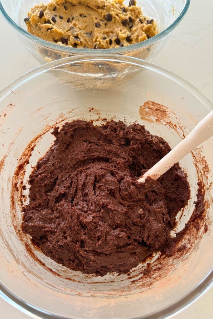 A bowl of sourdough brownie batter sitting on a white counter top with a bowl of sourdough chocolate chip cookie dough behind it.