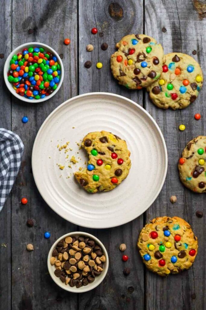 Sourdough monster cookies on wooden table with a cookie with a bite taken out sitting on plate next to a bowl of mini m&ms.