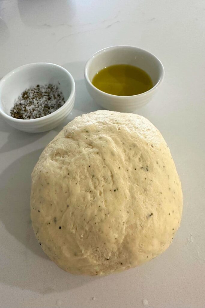 A ball of sourdough ranch cracker dough sitting on a counter top with two bowls of olive oil and dried herbs.