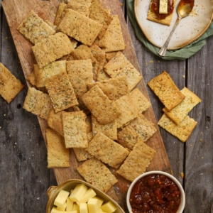 A tray of sourdough ranch crackers laid out on a rustic wooden board with a small dish of tomato chutney and some cheese cubes.