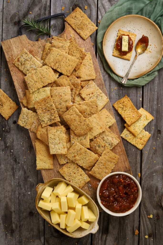 A tray of sourdough ranch crackers laid out on a rustic wooden board with a small dish of tomato chutney and some cheese cubes.