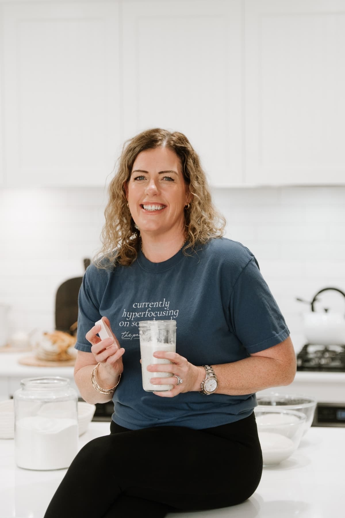 Kate Freebairn, founder of The Pantry Mama, holding a jar of sourdough starter. She is sitting on the edge of a white kitchen counter wearing a blue t-shirt and black pants.