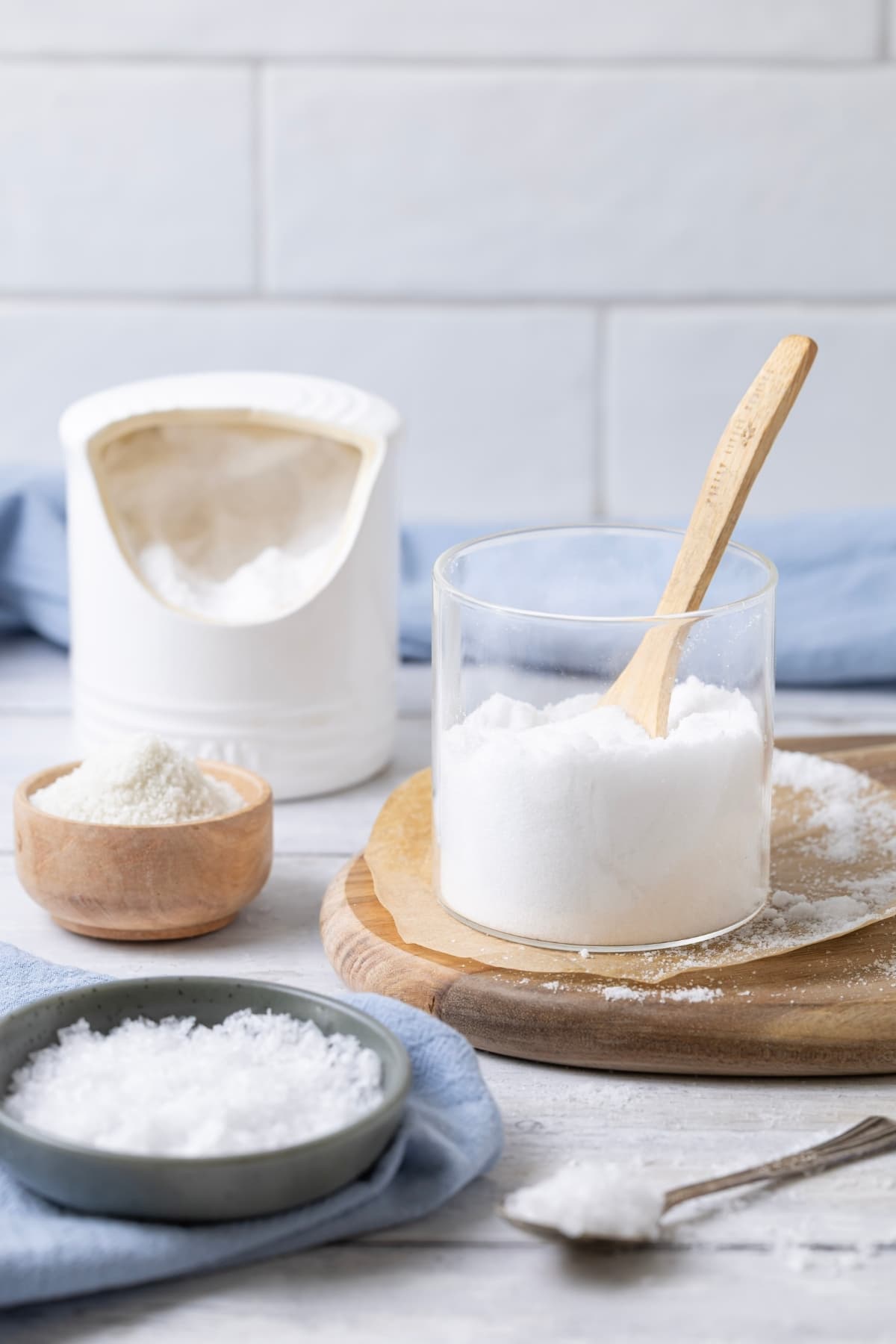 A photo showing different bowls of salt. There is a small bowl of flaky sea salt, a glass jar of fine sea salt and small wooden bowl of coarse salt. In the background there is a white salt pig.