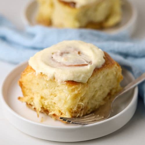 A sourdough cinnamon roll topped with vanilla cream cheese frosting sitting on a small white plate. There is a pale blue napkin the background of the photo.