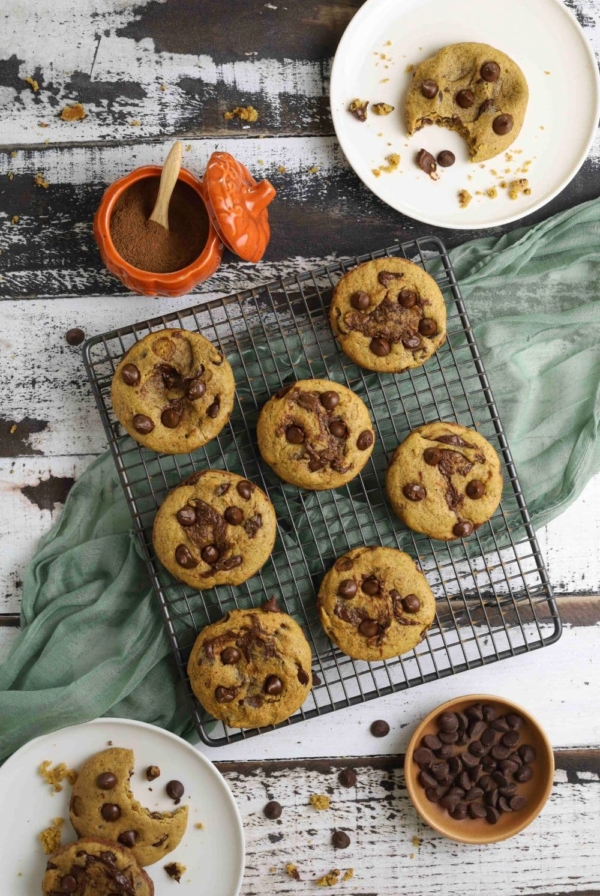 Sourdough pumpkin chocolate chip cookies on a cooling rack next to pumpkin spices and plates of partially eaten cookies.