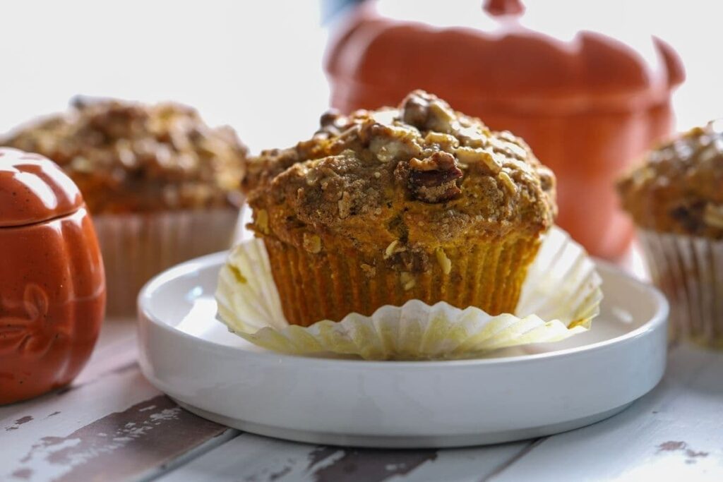 A sourdough pumpkin muffin sitting on a white plate, with the white wrapper pulled down. You can see an orange pumpkin in the background of the photo.