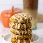 A stack of 5 sourdough pumpkin oatmeal cookies topped with maple glaze. They are on a white plate in front of a ceramic pumpkin and iced coffee.