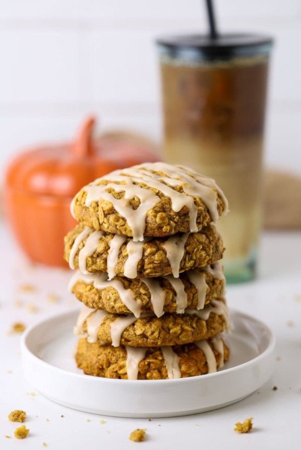 A stack of 5 sourdough pumpkin oatmeal cookies topped with maple glaze. They are on a white plate in front of a ceramic pumpkin and iced coffee.