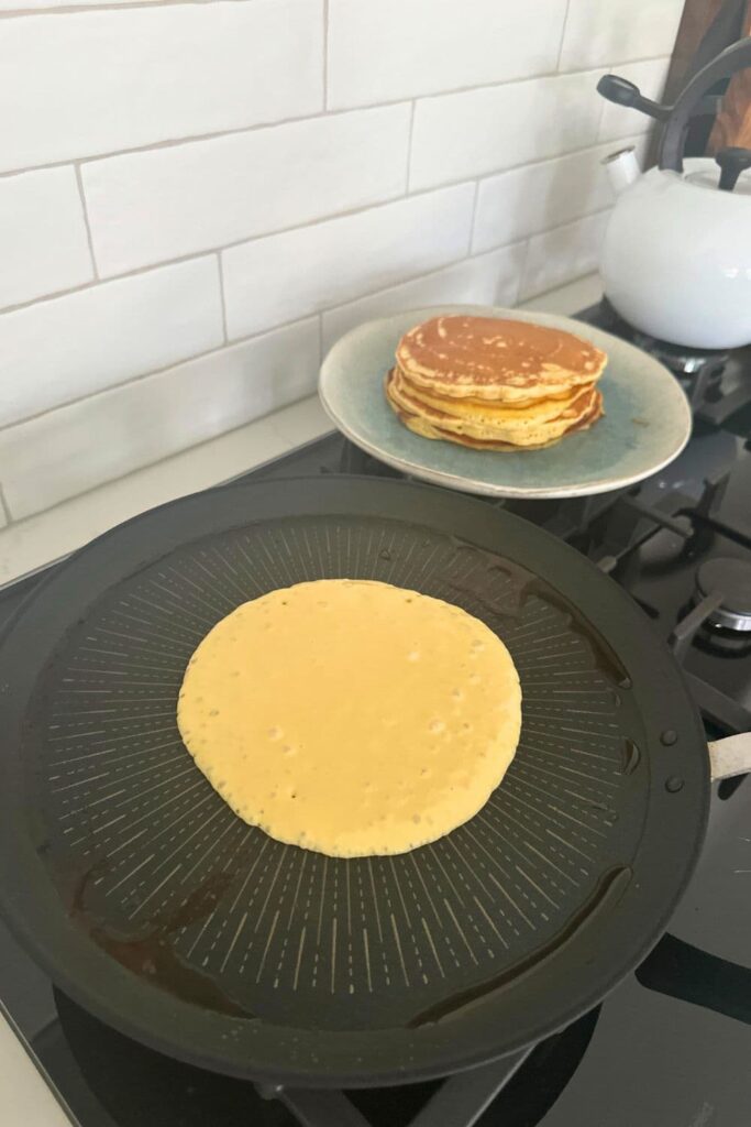 A sourdough pancake being cooked in a non stick crepe skillet. There is a stack of pancakes on a plate next to the skillet and a white kettle in the background of the photo.