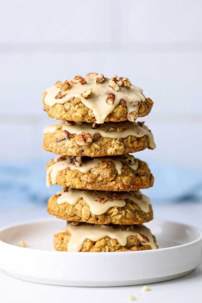 A stack of 5 maple pecan sourdough oatmeal cookies frosted with maple butter glaze sitting on a white plate. There is a pale blue napkin in the background.