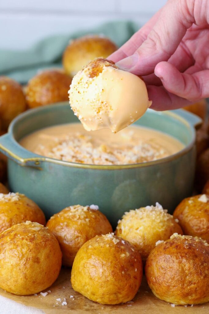 3 ingredient beer cheese dip served in a small green bowl. The bowl is surrounded by sourdough discard pretzel bites and one of the bites has been dipped into the beer cheese dip and is being suspended just above the bowl.