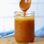 A glass mason jar filled with homemade caramel sauce being drizzled back into the jar with a spoon.